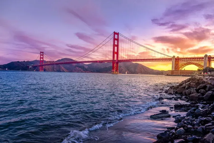 The Golden Gate Bridge at sunset with a multicolored sky and the San Francisco Bay in the foreground.